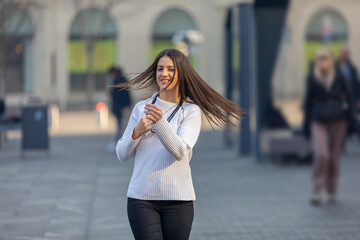 Fototapeta premium The concept of street fashion. Young beautiful model in the city. Beautiful teenager walking down the street. Hair fluttering in the wind. Excited beautiful girl expressing good emotions.