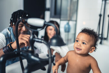 Mixed race toddler looking at shower head while standing in the bathtub. Parents watch their child develop. High quality photo