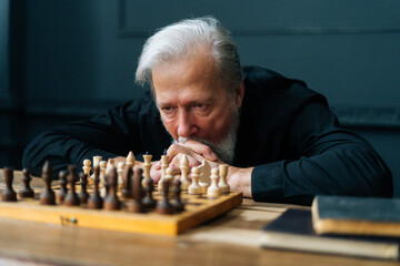 Close-up of pensive gray-haired senior older man thinking game strategy sitting on wooden table...