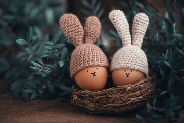 Two Easter eggs in crochet hats with rabbit ears in a nest on a wooden table. Soft focus.