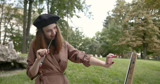 The girl artist in a black beret put the tip of the brush to her lips in the park