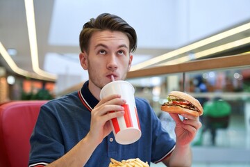 Portrait of young handsome man, guy is sitting on food court in shopping mall, eating, holding in...