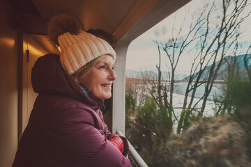 Woman (caucasian, 50s) wearing a puffer coat and warm hat, looking out a train viewing carriage 