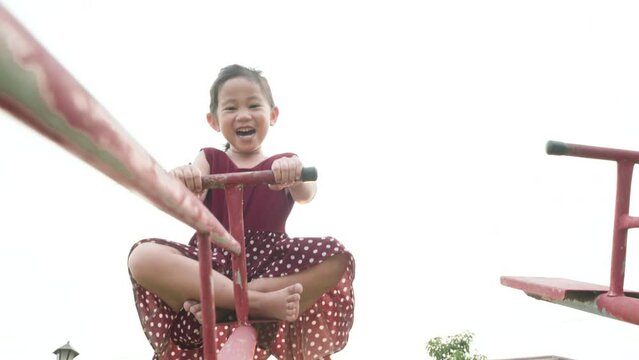 Happy smiling cute little girl playing on outdoor playground have fun with seesaw is up, freedom kid play chain teeter-totter or board swing under the sunset in summer