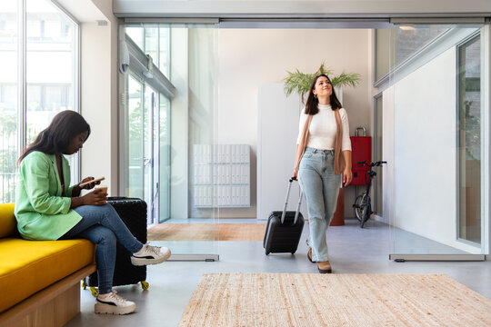 Happy young caucasian woman enters hotel reception with trolley suitcase. Copy space.