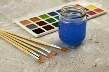 watercolor paints brushes and a jar of water on a gray textured surface