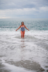 A plump woman in a bathing suit enters the water during the surf. Alone on the beach, Gray sky in the clouds, swimming in winter.