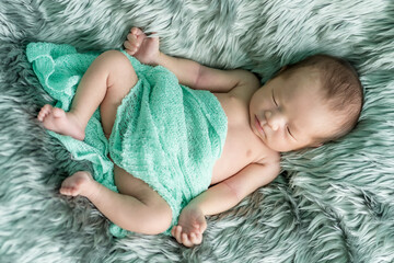 The newborn was wrapped in a green cloth with a headband and a bow was placed on a soft gray carpet. She is sleeping happily. Top view photo with soft focus.