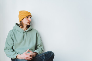 A hipster in a yellow hat sits with his hands folded together and looks away. Casual style in the clothes of a young man. A guy on a white background, an adult.