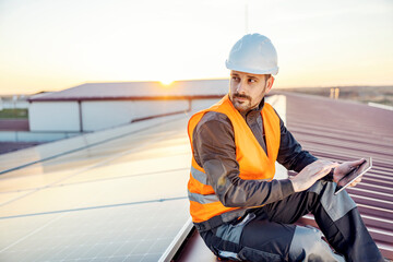 Worker sitting on the rooftop and using tablet to test solar panels at sunset.