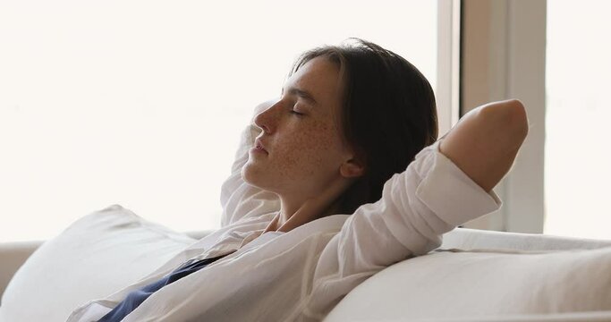 Happy calm young female student relax on cozy sofa with closed eyes breath deep enjoy cool humid air comfortable climate at home. Peaceful millennial woman chill on couch take break in vanity routine