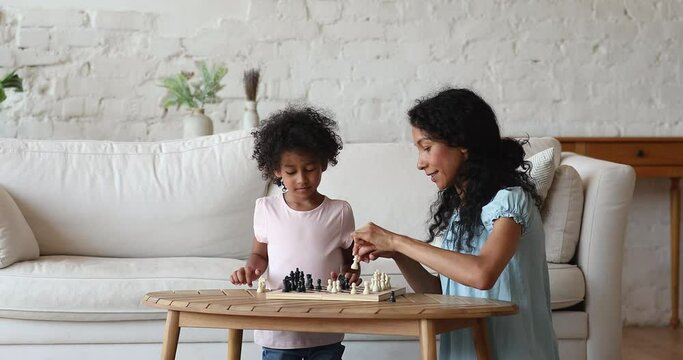 Caring young Black mother babysitter teach small daughter to play chess at living room. Grown elder sister compete with younger one in brain developing board game make move enjoy intellectual leisure