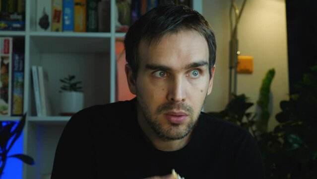 Close-up of a blue-eyed man watching horror on TV. Fear covers his mouth and eyes with his hand and turns away from the screen. A Caucasian man in black clothes eats popcorn while watching TV