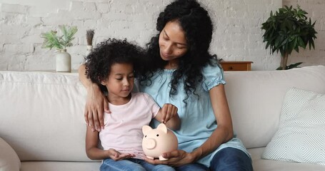 Young Black mother explain small daughter preschooler wise economy principle sit on sofa watch girl putting coins to funny piggybank. Caring mommy support child in wish to save pocket money for future
