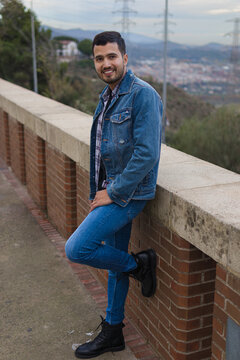 full length image of a hispanic guy dressed with blue clothes leaning on brick wall of a viewpoint with the city horizon behind him