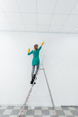 A young girl is engaged in repairs on the stairs and paints a white wall with a roller in a new apartment. Renovation of the interior and a new apartment. Housewarming and a desirable mortgage.