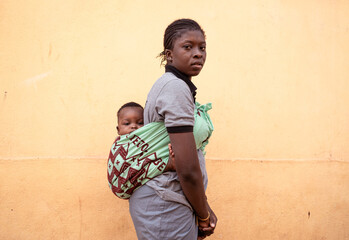 Young black African mother standing in front of a wall with her little baby on her back wrapped in...