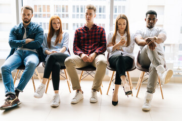 Front view of bored young diverse multi-ethnic job candidates waiting interview with hr, sitting in queue line row on chairs in modern office lobby on background of window and cityscape