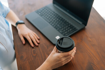 Close-up high-angle view of hands of unrecognizable young business woman sitting at desk with laptop in modern office room, holding cup with coffee. Tired businesswoman working at workplace.