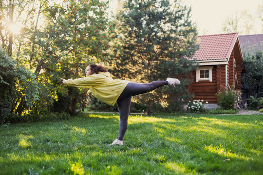 Caucasian fat woman standing on one hand doing yoga outside wearing sports clothes on green grass on backyard with country house and high trees in background. Body positive.