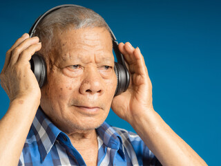 Senior man wearing wireless headphones to listen to a favorite song and looking away with a smile while standing on a blue background