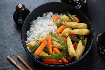 Middle closeup of vegetable stir-fry served over rice in a black bowl, studio shot on a dark-brown stone background