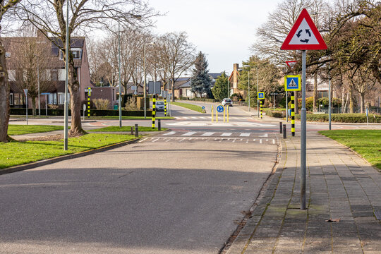 Triangular traffic sign for roundabout, pedestrian crossing, metal post with a triangular plate in red, black, white and arrows, roundabout and house in the blurred background, Netherlands