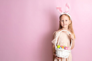 Happy Easter. A cute beautiful girl with bunny ears holds a basket of color eggs in her hands. Pink background. Space for text