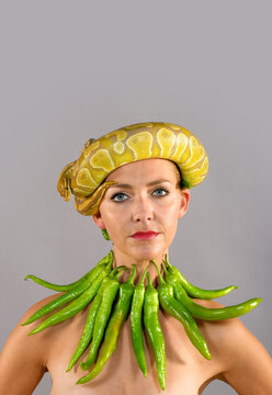 A beautiful woman wears a large green pepper
necklace while a yellow Python snake wraps 
itself around her head.
