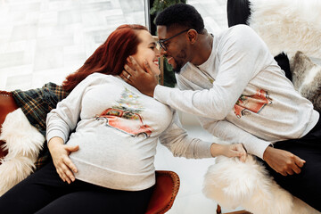 Adorable marriage couple sit near Christmas tree at home, caring African American husband hug beautiful pregnant wife, young family enjoy happiness, winter holidays concept