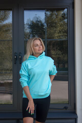 healthy activity. blonde girl in blue hoodie and black cycling shorts is standing casual near park cafe background and looking away with smile. sport lifestyle concept, free space