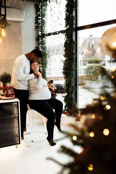 Happy marriage couple sit at the kitchen, prepare to Christmas dinner, overjoyed husband hug pregnant beautiful wife, young family expect for a baby, xmas celebration concept