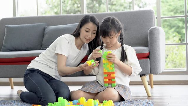 Happy Asian mom and little girl sitting on the floor playing with constructor blocks toy in living room at home. Family spend time together having fun with colorful plastic blocks