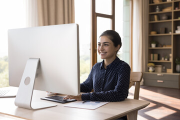 Smiling Indian female freelancer work on desktop pc at home office enjoy creative job. Glad young...
