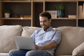 Smiling young man gamer relax on sofa enjoy new internet video game on laptop online at free leisure time. Happy positive millennial guy having fun using pc surfing social networks watching video blog