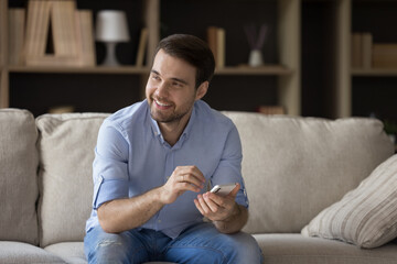 Positive guy sit on sofa hold phone look aside with smile enjoy good news received in online...
