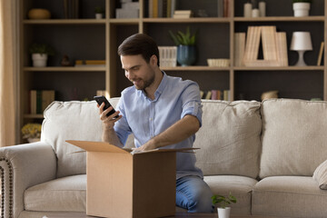 Excited guy blogger sit on couch unboxing postal package with consumer goods received from web shop by mail shoot video on mobile phone webcam. Happy young male open parcel delivery make photo on cell - obrazy, fototapety, plakaty