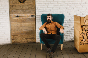 Successful bearded man in a turquoise chair on the terrace near a stack of firewood. Successful man of his country!