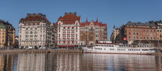 Naklejka premium Panorama view at the water front pier with apartment and hotel houses, commuting boats in in the calm water bay Ladugårdsviken a sunny winter day in Stockholm