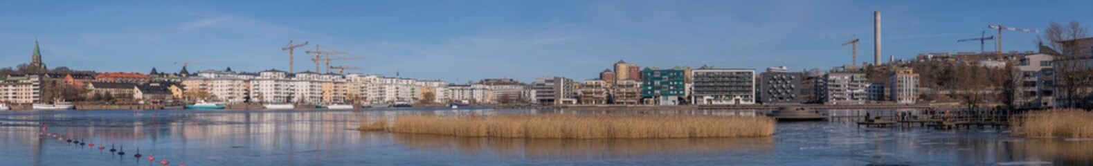 Fototapeta premium Panorama view at the icy bay Hammarby sjö with floes, apartment building and recreation area with commuting ferries a sunny winter day in Stockholm