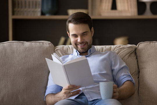 Smiling millennial guy spend cozy evening at living room sit on comfy couch read favorite paper book drink hot tea. Young man reader has pleasure enjoy classic novel fantastic story with cup of coffee