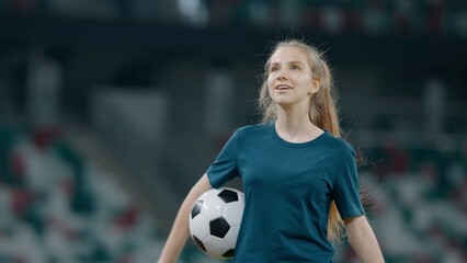 Portrait of Caucasian pre teen girl entering the field of huge soccer stadium, holding a ball,...