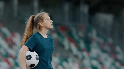 Portrait of Caucasian pre teen girl entering the field of huge soccer stadium, holding a ball,...