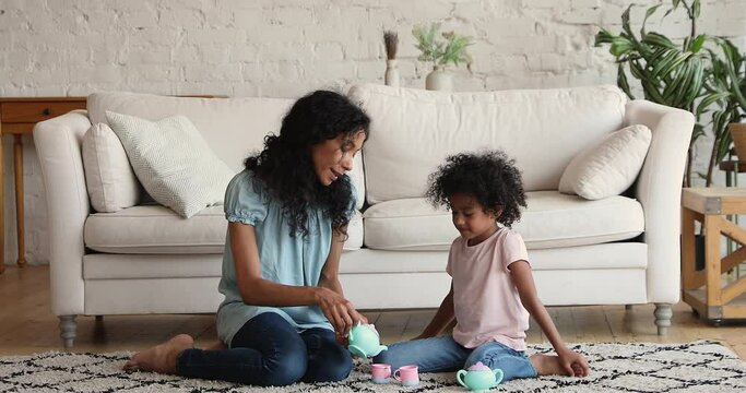 Happy Afro American foster mother adopted daughter best friends play on warm comfy carpet at living room imitate tea party. Happy nanny little kid having fun pretend drinking coffee using toy tea set