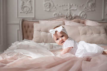 Obraz na płótnie Canvas a baby girl in a white headband and bodysuit crawls on a bed in a room with a beautiful interior