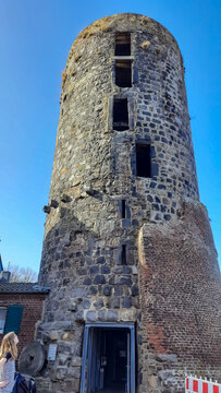 M&uuml;hlenturm Schloss Liedberg. Alter historischer steinerner Turm in einem deutschen Dorf. &Uuml;berbleibsel einer alten Burg oder Stadtmauer. Eine Frau mit Maske steht am Fu&szlig;e des Gem&auml;uers schaut nach oben.