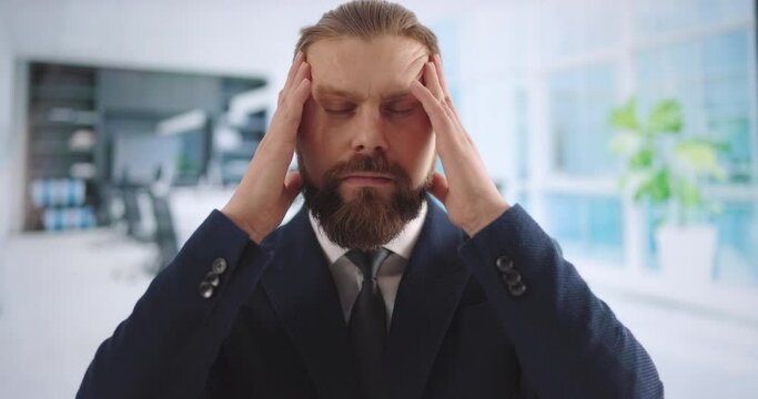 Tired caucasian businessman in suit massaging temples while suffering from headache during work at office. Overwork and stress concept.