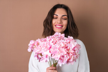 A young woman with wavy voluminous hair on a beige background with bright pink lipstick lip gloss in a white sweater holds a bouquet of pink flowers