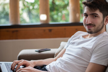 jeune homme qui travaille avec son ordinateur portable, allongé dans un fauteuil. Il travaille à la maison, en télé travail. Il est relax, zen et détendu