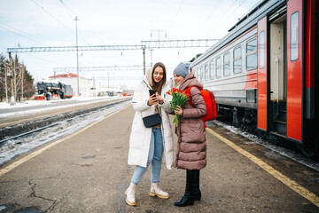 two girls on the station platform with flowers happily looking into a smartphone - obrazy, fototapety, plakaty